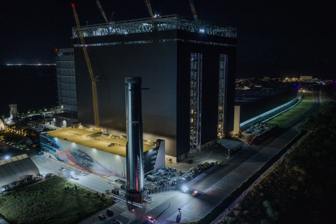 An aerial nighttime view shows a massive building under construction with cranes on top, illuminated lights, and a SpaceX Starship rocket booster standing vertically next to it.