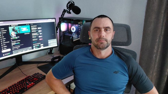 A man in a blue athletic shirt sits in a gaming chair in front of a computer setup with a curved monitor, RGB-lit PC tower, and red backlit keyboard, with chat/messaging visible on the screen.