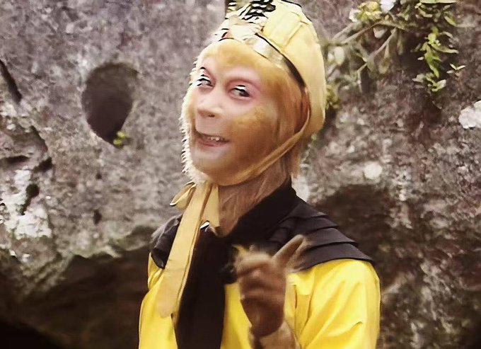 An actor in a yellow monkey costume with a flesh-toned monkey face mask and a Chinese character on the headpiece, smiling with pointed teeth against a rocky cave background, appearing to be a scene from a classic Chinese television adaptation of Journey to the West.