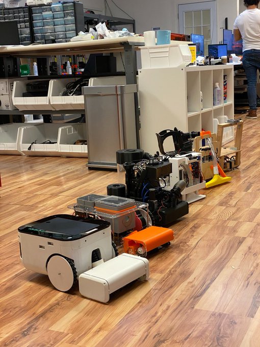A robotics workshop or maker space with various robots and mechanical projects lined up on a wooden floor, including a white wheeled robot in the foreground.