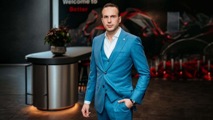 A man in a sharp blue three-piece suit stands confidently in a sleek modern lounge, with a "Welcome to Better" sign visible behind him alongside a racing-themed mural. The professional setting suggests a business or media interview context, aligning with the post's theme of discussing regulatory developments like MiCA, the Clarity Act, and the UAE's crypto framework shaping industry adoption.
