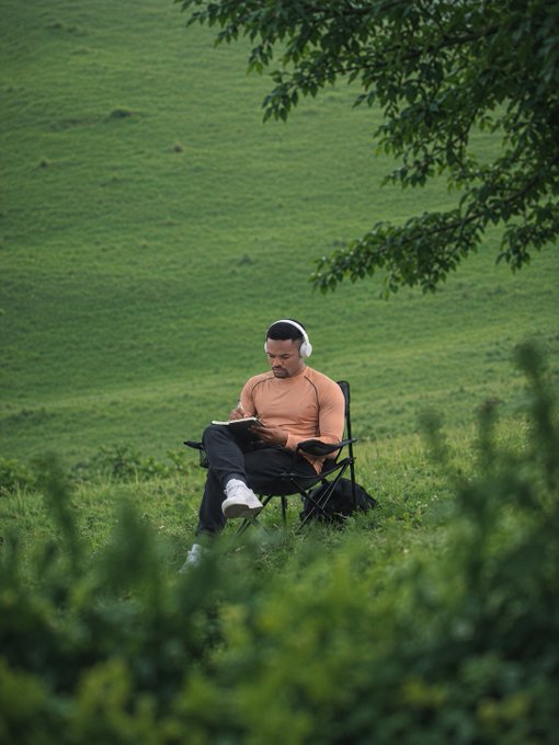A man wearing an orange long-sleeve shirt and white headphones sitting in a folding chair on a grassy hillside, writing in a notebook surrounded by green foliage.