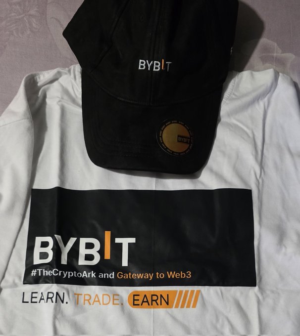 The image features a T-shirt with the words "Bybit" written across it along with other information related to Bitcoin and web transactions, such as learning, trade, earn, and others. There is also an ear tag attached to the shirt, which contains a small amount of money. Alongside this, there is a baseball cap displayed in the image that appears to be black or dark-colored. The arrangement of these items suggests a connection between the Bitcoin merchandise package and the hat, perhaps indicating some affiliation or partnership with the brand.