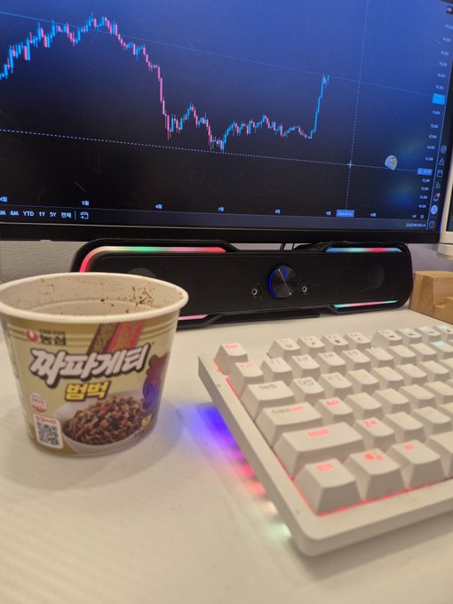 A desk setup showing a Bitcoin trading chart on the monitor alongside a cup of "짜파게티 알약" (Jjapagetti Al-yak) instant noodles and a white RGB mechanical keyboard. The image matches the post's message about preparing cup noodles after noticing Bitcoin starting to move right before going to bed.