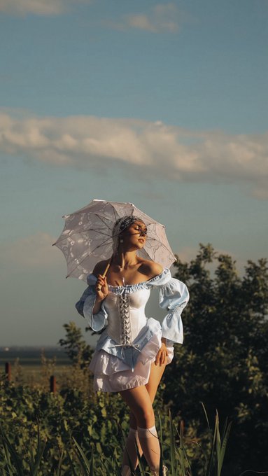 A woman in an elegant white off-shoulder ruffled dress stands in a grassy field holding a white lace parasol, with a scenic landscape and blue cloudy sky behind her.