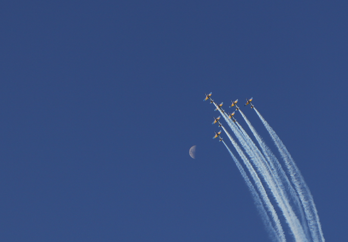 A formation of aircraft, appearing to be the Blue Angels, flying in a diagonal line against a deep blue sky, leaving white contrails behind them near a visible crescent moon.