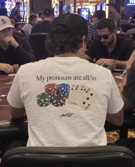A poker player wears a white t-shirt featuring the pun "My pronouns are all/in" alongside images of poker chips and cards (7, 8, 5, 2, 3), visually tying into the giveaway post's likely theme of gambling or crypto poker. The shirt cleverly blends poker terminology with current pronoun discourse, suggesting the giveaway may target poker or gambling enthusiasts.