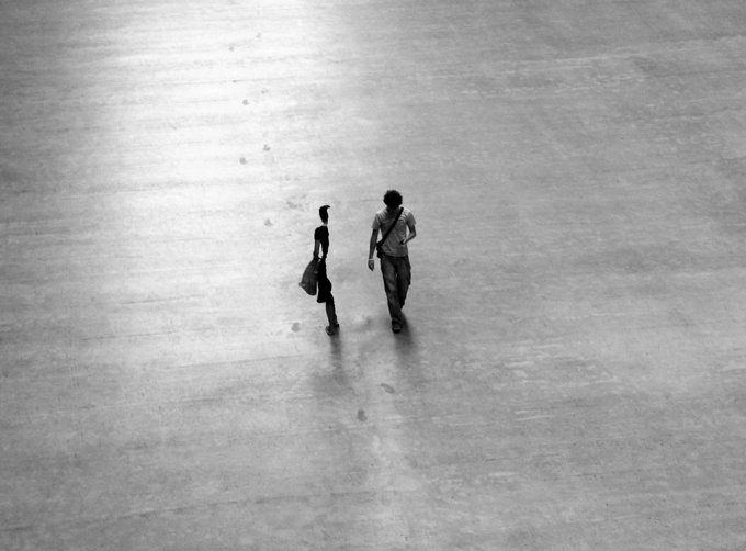 A black and white photograph captures two people from behind walking across a vast, empty concrete floor, leaving a trail of footprints.