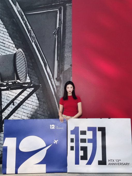 A woman in a red t-shirt stands behind two banners reading '12' and '捌' (eight in Chinese), promoting the 'HTX 13th ANNIVERSARY'.