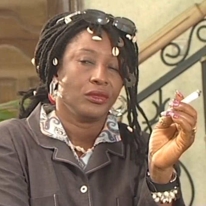 A woman with long braided hair adorned with cowrie shells and sunglasses on her head smokes a cigarette while posing near a staircase, fitting the "Carry belle" (smoking) option from the post's list of ways to scatter one's life at a young age.