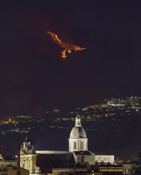 Nighttime photograph showing a glowing volcanic eruption or lava flow on a mountainside above a hilltop town with a prominent domed cathedral in the foreground.