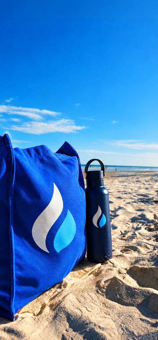 A blue canvas bag and matching black water bottle, both branded with a white and blue droplet/flame logo, sit in the sand of a sunny beach. The matching branded gear visually "brings the alpha to the beach," symbolizing bold, branded confidence in a laid-back environment.