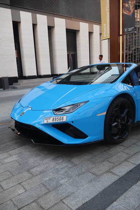 A bright blue Lamborghini Huracán with Dubai license plate "D 89566" is parked outside a building displaying "Where Collection Flow" signage. The "GM if you GM" caption playfully suggests greeting the luxury supercar with the same energy it commands, as if saying "good morning" only to those deemed worthy of its presence.