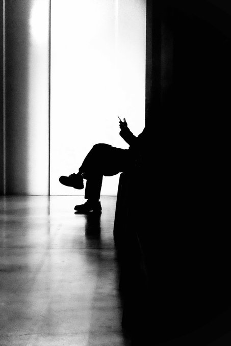 A silhouette of a figure seated in a chair, absorbed in their phone, is dramatically backlit against a bright doorway, embodying the contemplative essence of being "in the shadows" while a piece of light (the screen) draws focus. The high-contrast black-and-white composition visually echoes the post's themes of introspection and the interplay between darkness and illumination.