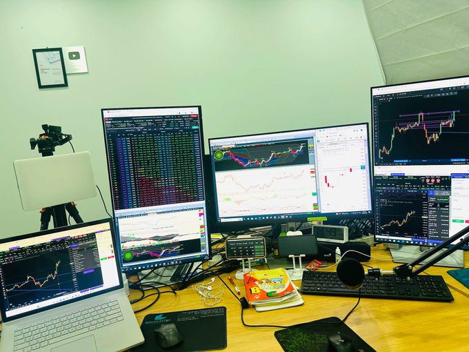 A trading desk setup featuring multiple monitors displaying financial charts and stock data, a laptop showing trading software, a microphone, and snack packages, indicating an active trading or crypto analysis workspace.