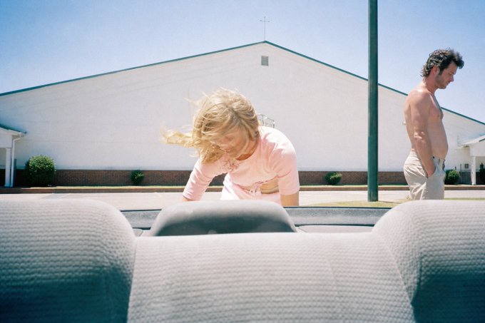 Photo taken from inside a car looking out at a blonde woman in a pink shirt leaning into the trunk area, while a shirtless man in light pants stands nearby in front of a white church-like building.