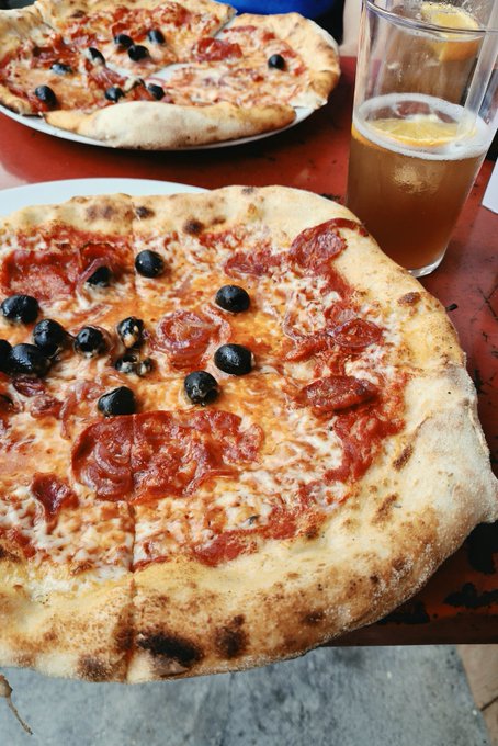 A pizza topped with pepperoni and black olives sits on a red table, accompanied by a glass of beer and a second pizza partially visible in the background.