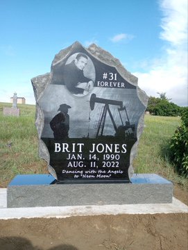 A granite memorial headstone engraved with '#31 FOREVER BRIT JONES JAN. 14, 1990 AUG. 11, 2022' and a portrait, dedicated 'In Loving Memory with Pride & Dignity' by 'Mom-Mom', featuring a bass guitar image.