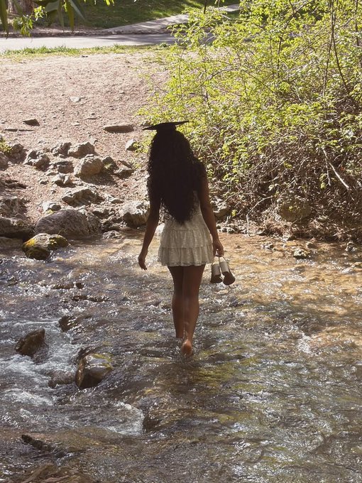 A woman in a white dress walks barefoot through a shallow rocky stream, carrying boots in one hand with long dark hair flowing down her back.