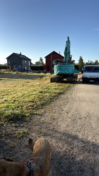 A dog watches as a teal Kobelco excavator works near three houses under a clear sky, with a post referencing an "IRL launchpad" deployment of three (soon four) houses and asking whether to shill "ARC." The image visually mirrors the "houses deployed" theme, showing the construction equipment and partially built neighborhood that the post humorously likens to a crypto project launch.