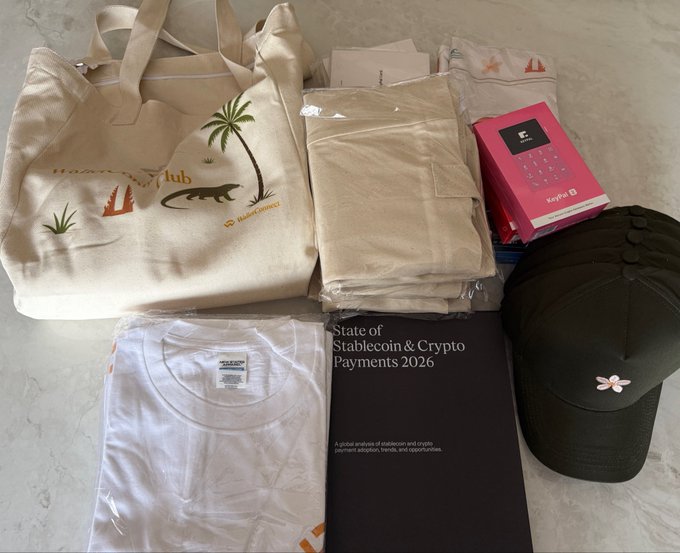 A flat-lay of promotional merchandise including a Hub tote bag with a palm tree design, a white t-shirt, a dark cap with a flower logo, a report titled 'State of Stablecoin & Crypto Payments 2026,' and a KeyPal device in pink packaging.