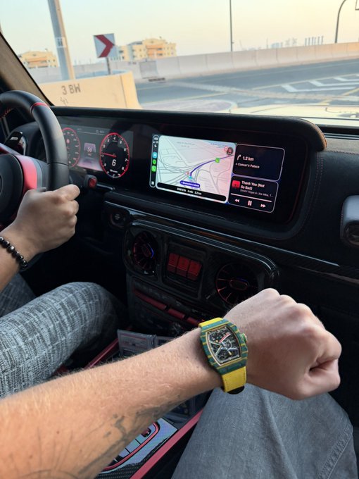 A driver's wrist flexes to show off a yellow watch in the foreground, with the car's dashboard displaying Apple CarPlay navigation set for 1.2 km to "Al Garhoud Bridge" and music playing from "Spoti-Top Shar" — a relaxed morning cruise vibe.