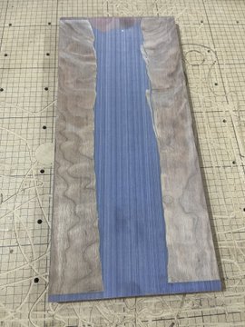 A rectangular piece of wood with a blue-painted river-like design running down the center, bordered by natural gray wood grain, sits on a grid mat.
