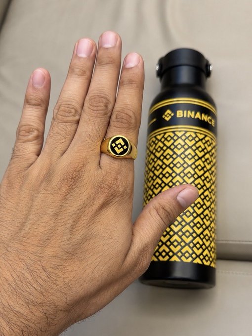 A man's hand displays a gold Binance ring next to a black and gold patterned Binance water bottle.
