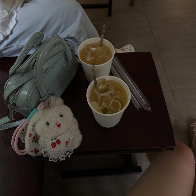 The image features a wooden table with two cups of tea sitting on top of it. One cup has ice cubes in it, while the other does not. A stuffed animal, possibly a rabbit or a teddy bear, is placed next to one of the cups. The scene appears to be set in an office environment, as there are chairs and a handbag visible around the table.