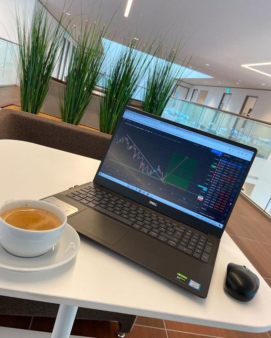 A Dell laptop on a white table displaying a financial candlestick chart, accompanied by a cup of espresso and a black wireless mouse in what appears to be a modern indoor lounge or cafe.