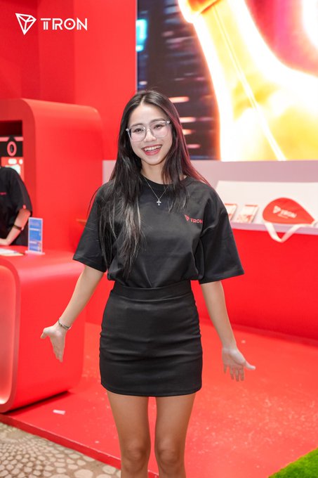The image shows a smiling woman with glasses wearing a black TRON-branded t-shirt and skirt, posing at the TRON booth at what appears to be a blockchain event called Conviction, matching the post's invitation to visit the booth.
