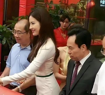 A photo of a woman in a white dress with a green belt cutting a ribbon at an event, surrounded by men in suits and other attendees, with Chinese characters visible in the background.