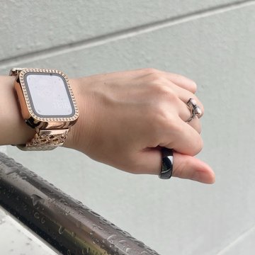 A wrist displaying a gold Apple Watch with a decorative bezel and floral band, alongside a black ring on the middle finger and a rose-gold ring on another finger.