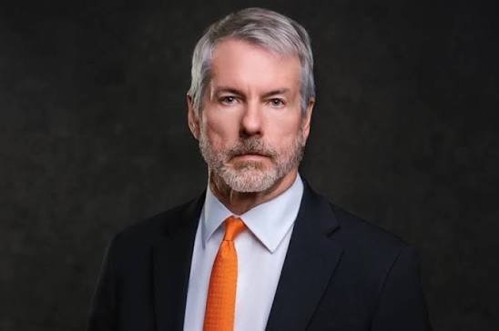 A formal portrait photograph of an older man with gray hair and beard wearing a black suit, white shirt, and orange tie against a dark background.