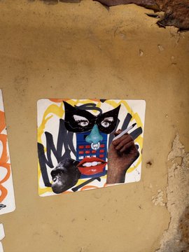 The image features a sticker with an abstract design of a face, including a mask, holding a cigarette between its teeth. The sticker is attached to a wall or surface using tape. Below the sticker, there's text that reads "Started from the bottom now we're here". This suggests that the sticker was created by someone who started at the bottom and worked their way up to the top of the image, possibly as part of an art project or personal expression.

The image also contains a cell phone with a picture of a monkey on it, which adds another layer of complexity to the overall composition. The combination of these elements creates a visually interesting and thought-provoking piece that invites viewers to explore its meaning and context further.