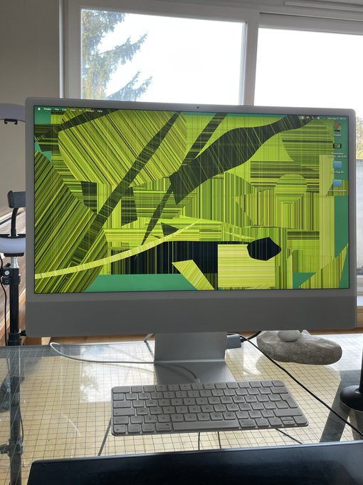 An iMac desktop displays a distorted green and black abstract wallpaper with visual glitches on its screen, accompanied by a wireless keyboard on a glass desk.
