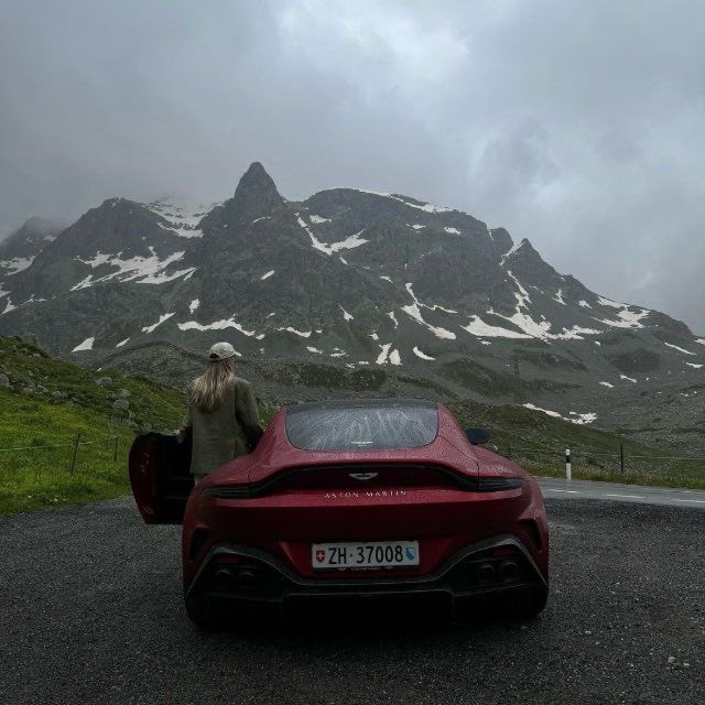 A red Aston Martin sports car parked on a mountain road with a person standing by the open door, set against a dramatic backdrop of snow-capped peaks under cloudy skies; the Swiss license plate reads 'ZH-37008'.