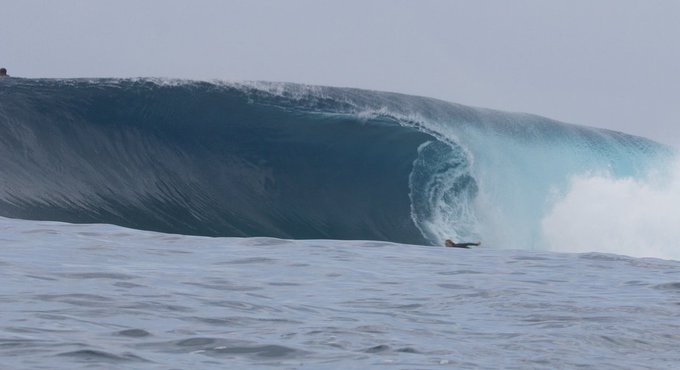 A lone surfer, dwarfed by the towering wave, sets up for the barrel beneath a massive, curling crest of blue-green water while white foam explodes to the right. The scene captures the thrilling moment of commitment, where focus and awareness of the wave's peril are essential to survive the ride.