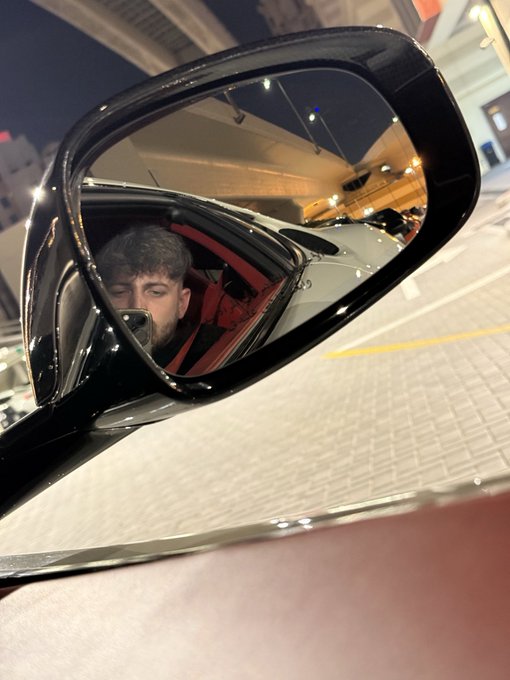 A nighttime view captured through a car's side mirror, reflecting the driver holding a phone, with a red sports car visible in the background on the street.