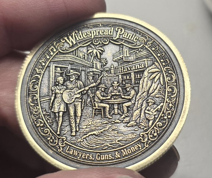A close-up of a commemorative coin held between fingers, engraved with 'Widespread Panic', 'Lawyers, Guns, & Money', and a detailed scene featuring 'Havana' in the background.