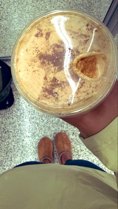 A first-person view looking down at a plastic cup containing a creamy, cinnamon-dusted beverage and a small cookie, held above brown suede moccasin shoes on a tiled floor.