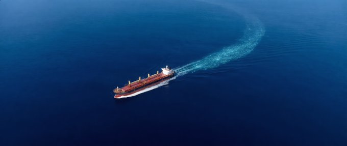 Aerial photograph of a red and white cargo ship sailing through deep blue ocean waters, leaving a curved white wake behind it.