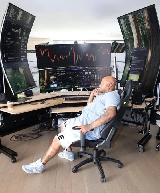 A bald, tattooed man in shorts sits in a gaming chair looking at a multi-monitor trading setup displaying candlestick charts, with a large central screen showing red bearish price action.