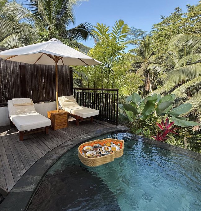 A floating breakfast tray serves assorted dishes, including fruit and small bowls, across an infinity pool surrounded by tropical foliage and cushioned lounge chairs under a white umbrella. The post "Who's active on the weekends" pairs perfectly, as the leisurely poolside morning ritual suggests an active and indulgent weekend escape.