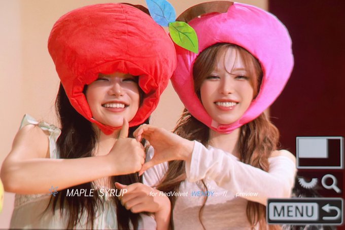 Two smiling women wearing large plush fruit-shaped hats (a red strawberry and a pink peach with leaves) pose together making finger hearts, with text reading 'MAPLE SYRUP for Red Velvet WEEKLY preview' visible.
