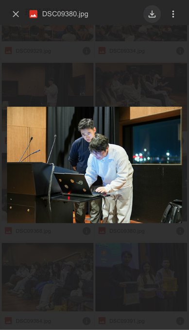 A screenshot of a photo gallery app showing an image of two men working at a presentation podium, with thumbnails of other event photos visible above and below.