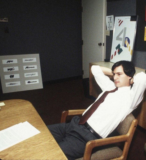 A vintage photograph shows a young man in a white shirt and tie leaning back in a chair in an office setting.