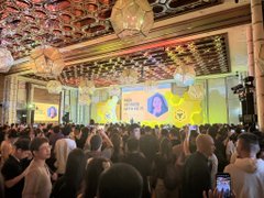 A packed cryptocurrency conference hall features an elaborate ceiling with glowing geometric lights above a stage displaying the Binance logo, surrounded by attendees taking photos and recording the event, capturing the energy described in the caption about being part of Binance's vibrant offline crypto community.