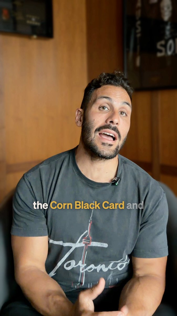 Man with a beard wearing a gray Toronto t-shirt sitting in a chair and gesturing while talking, with on-screen text reading 'the Corn Black Card and'.