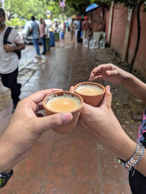 Two hands clinking small clay cups (kulhads) of chai tea together on a wet sidewalk street scene with pedestrians and water jugs in the background.